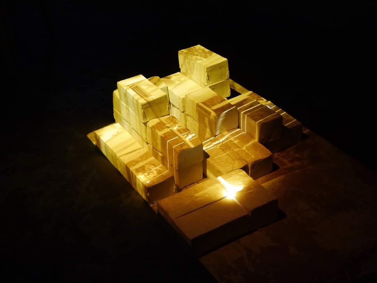 Cast ceramic blocks arranged on a dark floor and lit from within, glowing pale gold against the black.