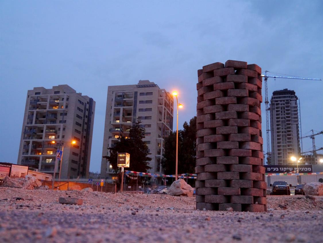 A column of stacked cement bricks standing on broken ground at dusk, with new apartment towers and a crane behind it.