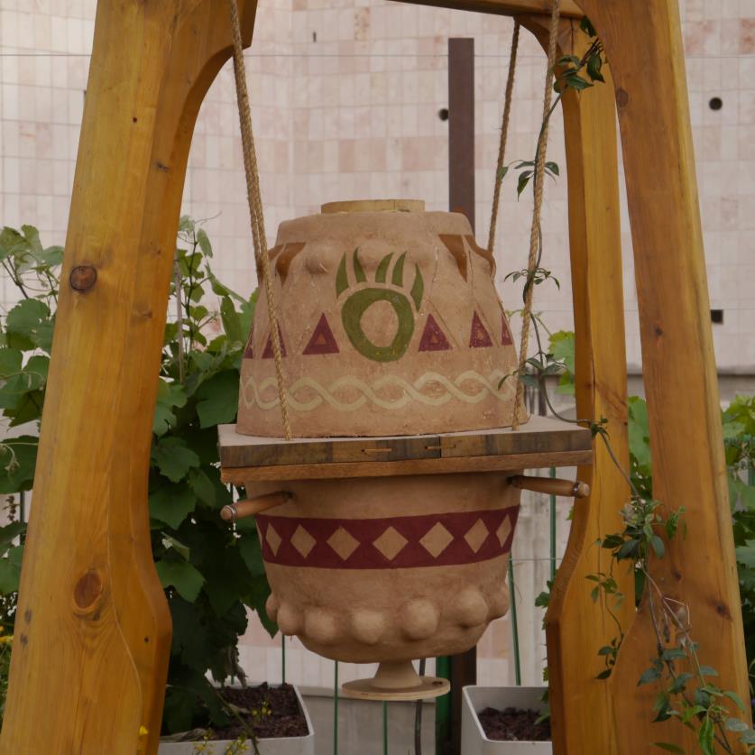 A patterned earthen beehive vessel hung inside a peaked wooden frame, climbing plants around its base.