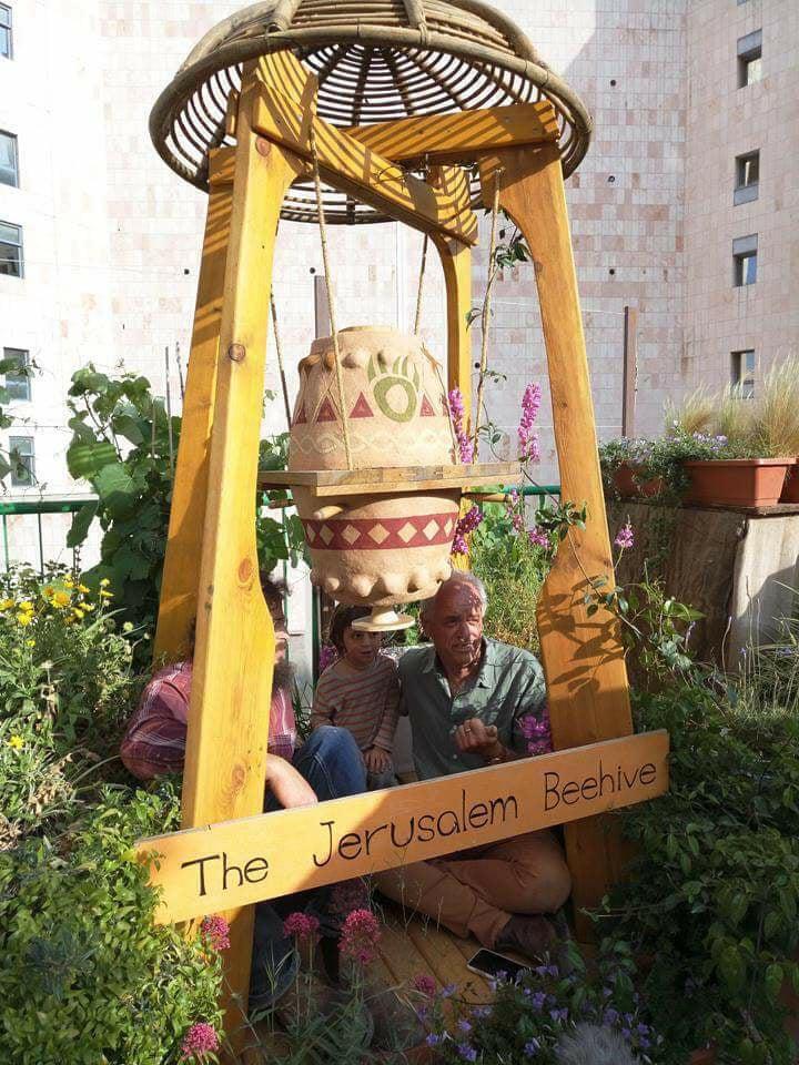 The beehive in its rooftop garden behind a carved wooden sign reading The Jerusalem Beehive, with people sitting beyond it.