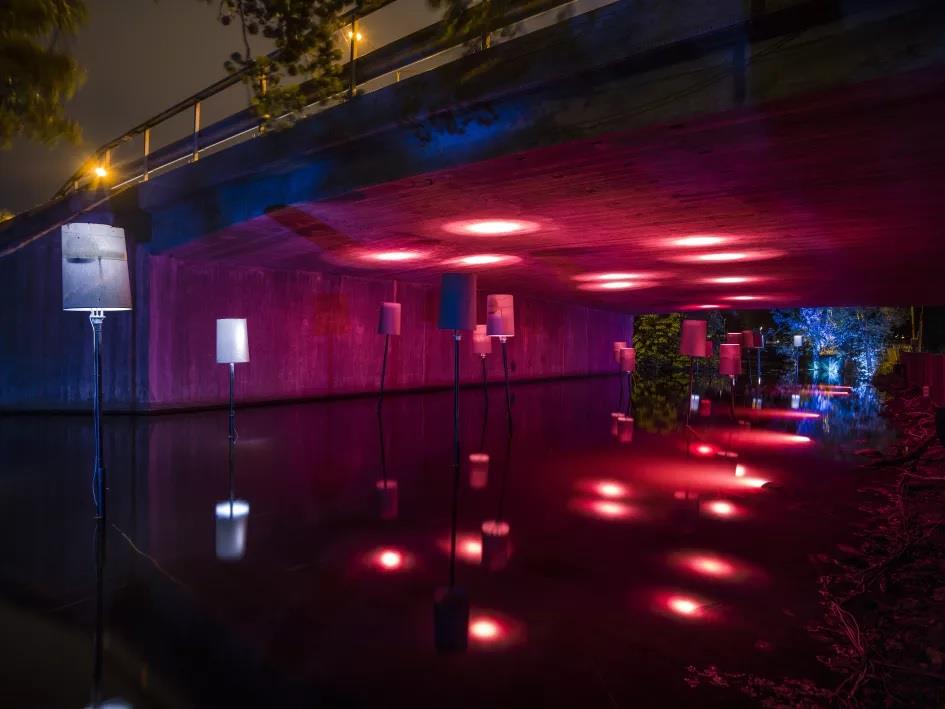 A concrete underpass lit deep red, with tall standing lamps in shallow water and their reflections running back into the dark.