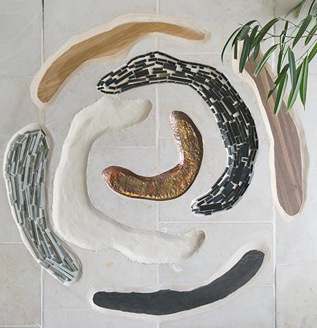 A spiral relief set into a pale wall, its arcs made of copper, dark stone, wood and woven fabric.