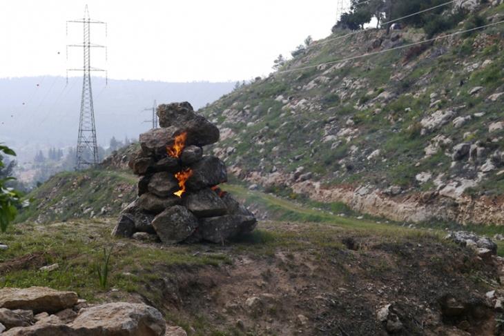 A cairn of stacked stones burning on a bare hillside in daylight, an electricity pylon and hills behind.