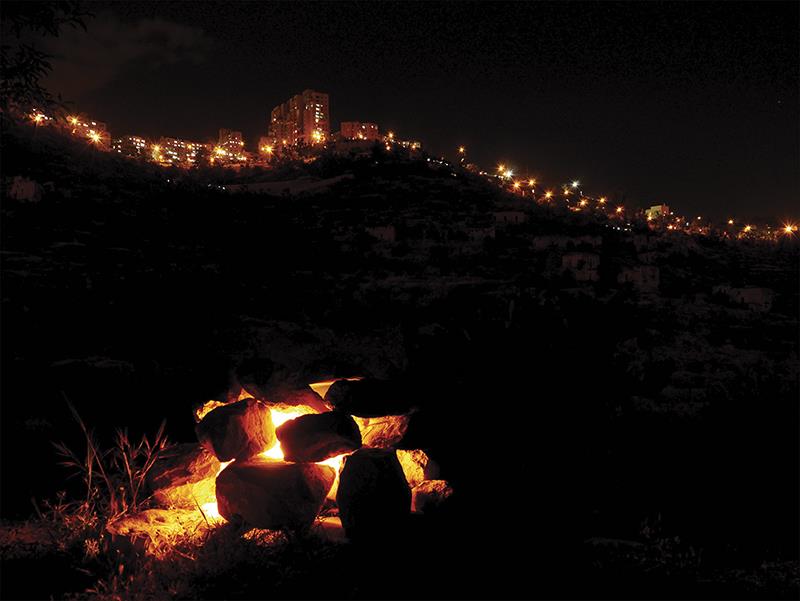The same cairn burning at night, its stones glowing from within, the lights of the city strung along the ridge above.